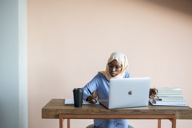 Female university student in a hijab working at a laptop alongside a pile of textbooks