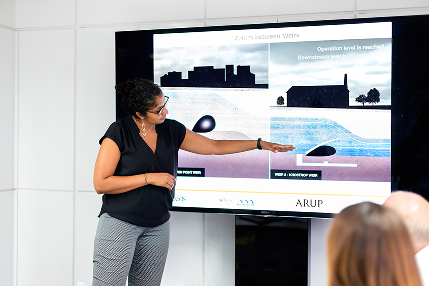 Photographt of woman presenting to a room a slideshow of scientific diagrams on a big screen.