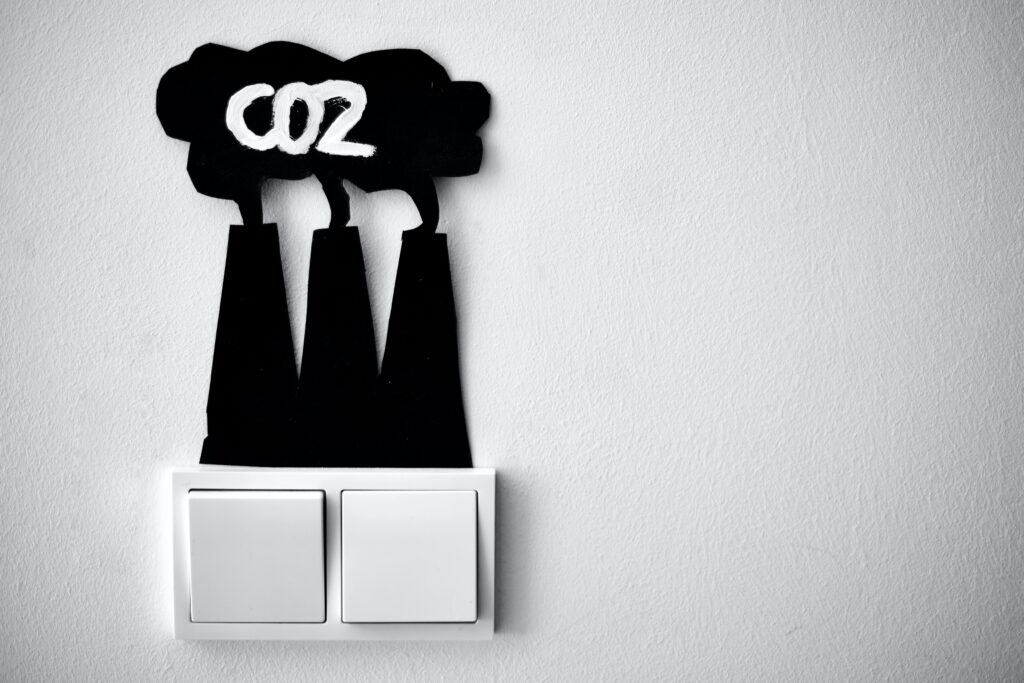 A light switch with two switches - one is off and one is on. Coming out of the top of the switches is a black cut-out sillhouette of factory smokestacks with billowing smoke. CO2 is written in the cloud of smoke.